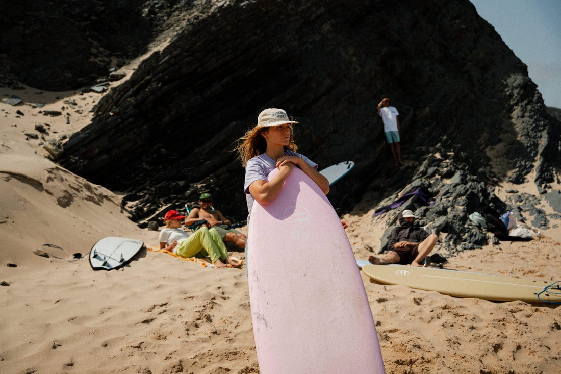 Surfista con tabla rosa Fly en la playa, rodeada de amigos disfrutando de un día de surf.