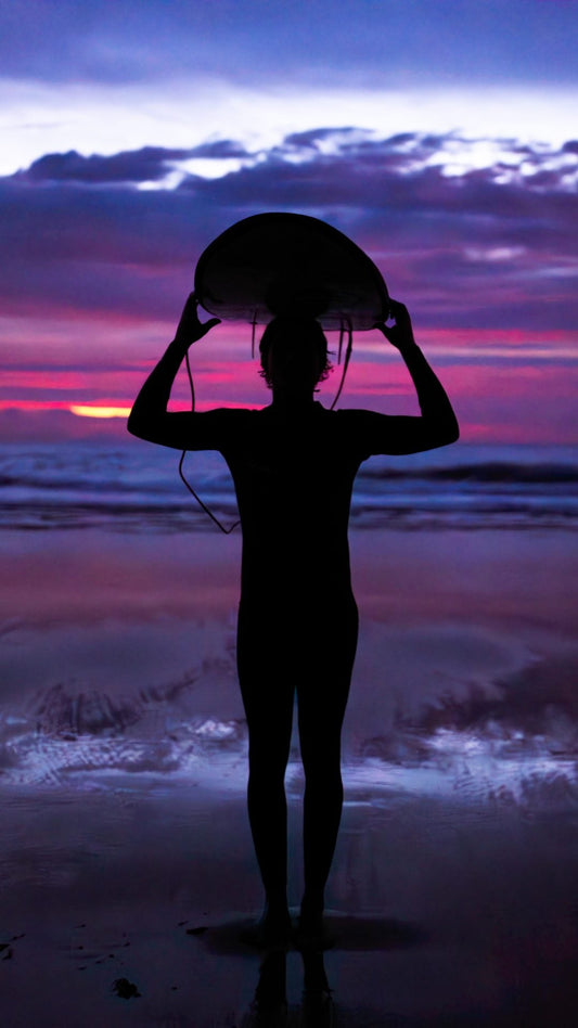 Surfista al atardecer sosteniendo su tabla frente al mar y pensando en la Recuperación después de surfear