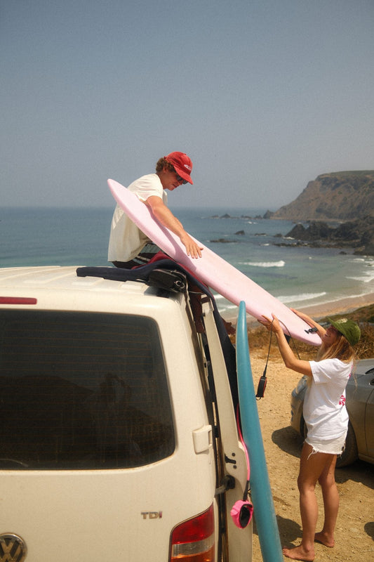 Dos surfistas preparan sus tablas de surf junto a una furgoneta frente al mar durante un road trip de surf en otoño, en un paisaje costero.