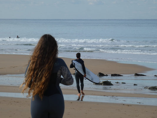 Dos surfistas entrando al agua con neoprenos en una playa, listos para disfrutar de las olas.