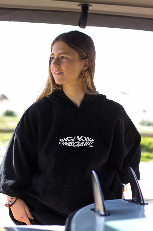 Persona con un poncho negro Fly junto a una tabla de surf, en un ambiente exterior luminoso.