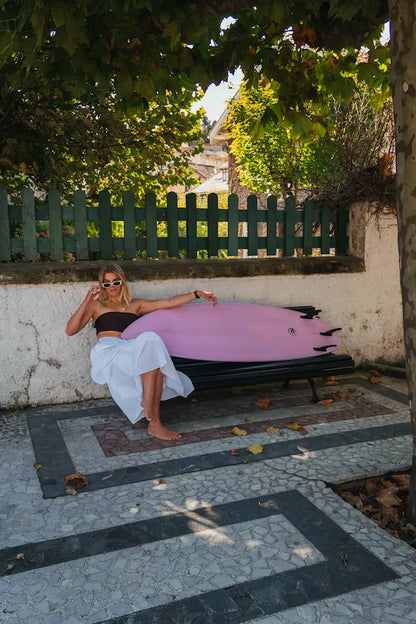 Chica en un banco posando con la tabla de surf rosa de fibra.