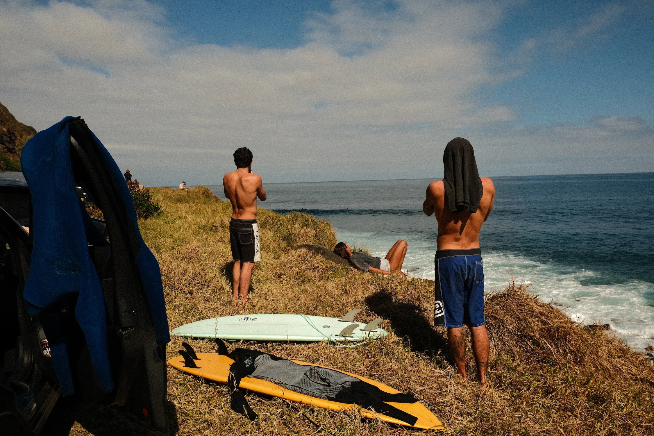 Dos surfistas de pie en un acantilado de hierba observando el mar junto a sus tablas de surf y un coche.